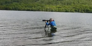 Photographer Rob Clifford standing waist-deep in Coniston Water with a telephoto lens on a tripod during the Bluebird K7 Festival, May 2026