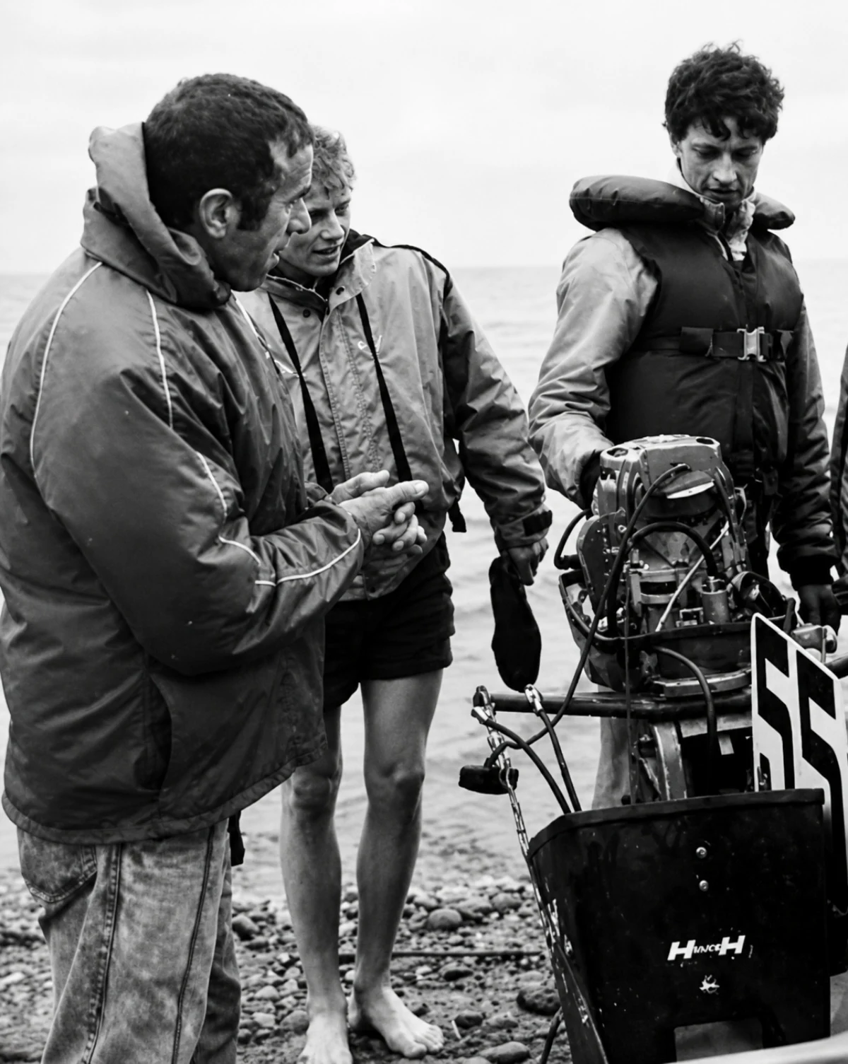 Valdemaras Petrūnas discussing engine setup with athletes on a rocky beach, vintage black and white photograph