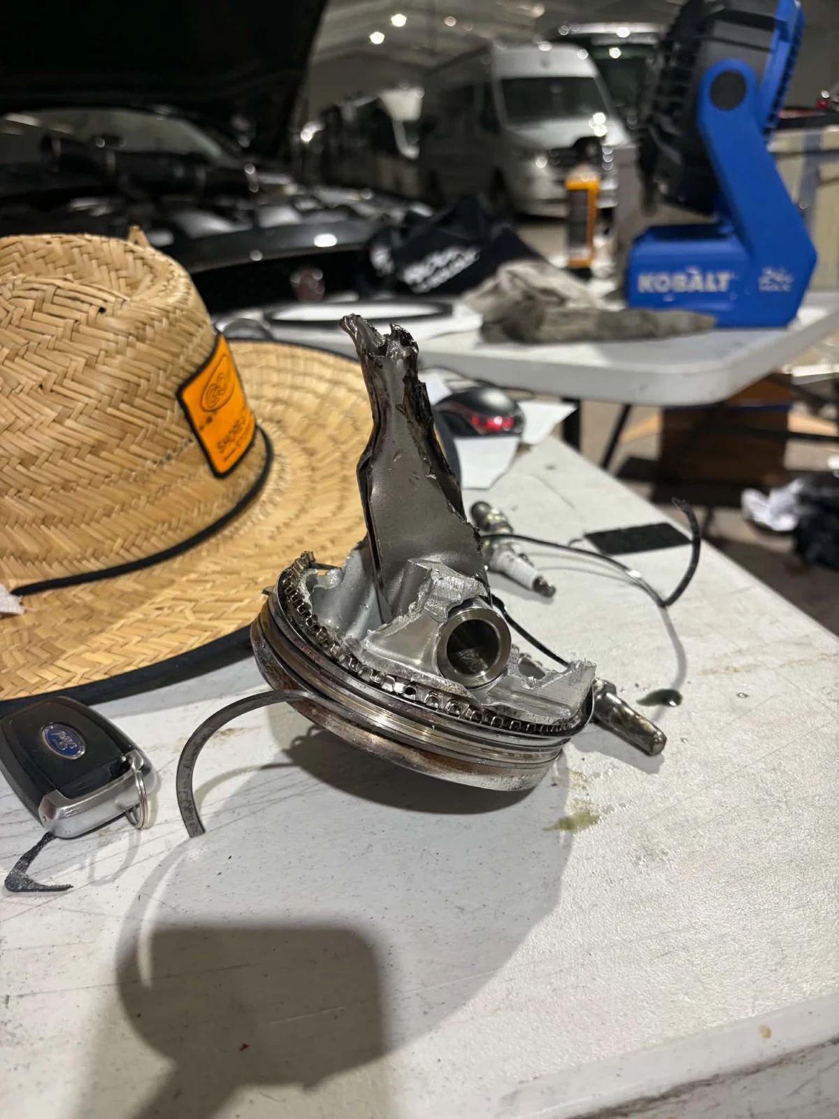 Failed piston with a broken connecting rod torn through it, on a workbench beside a straw hat and car key