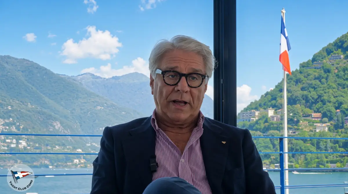 Vincenzo Iaconianni in a dark jacket and glasses, speaking in front of a window overlooking Lake Como