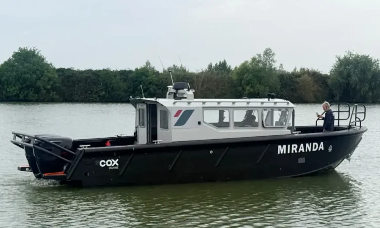 Cox Marine test and development vessel Miranda, a black Tideman RBC1200 HDPE workboat with white wheelhouse and twin Cox Marine V8 diesel outboards, under way on a river